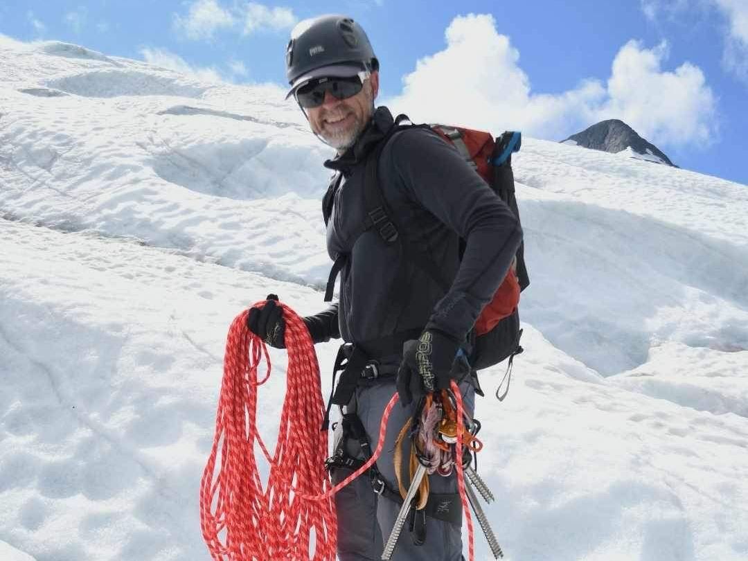 A person with a helmet and backpack stands on a snow-covered glacier with climbing equipment and a red rope against the blue sky.