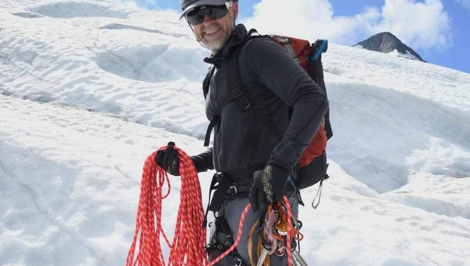 A person with a helmet and backpack stands on a snow-covered glacier with climbing equipment and a red rope against the blue sky.