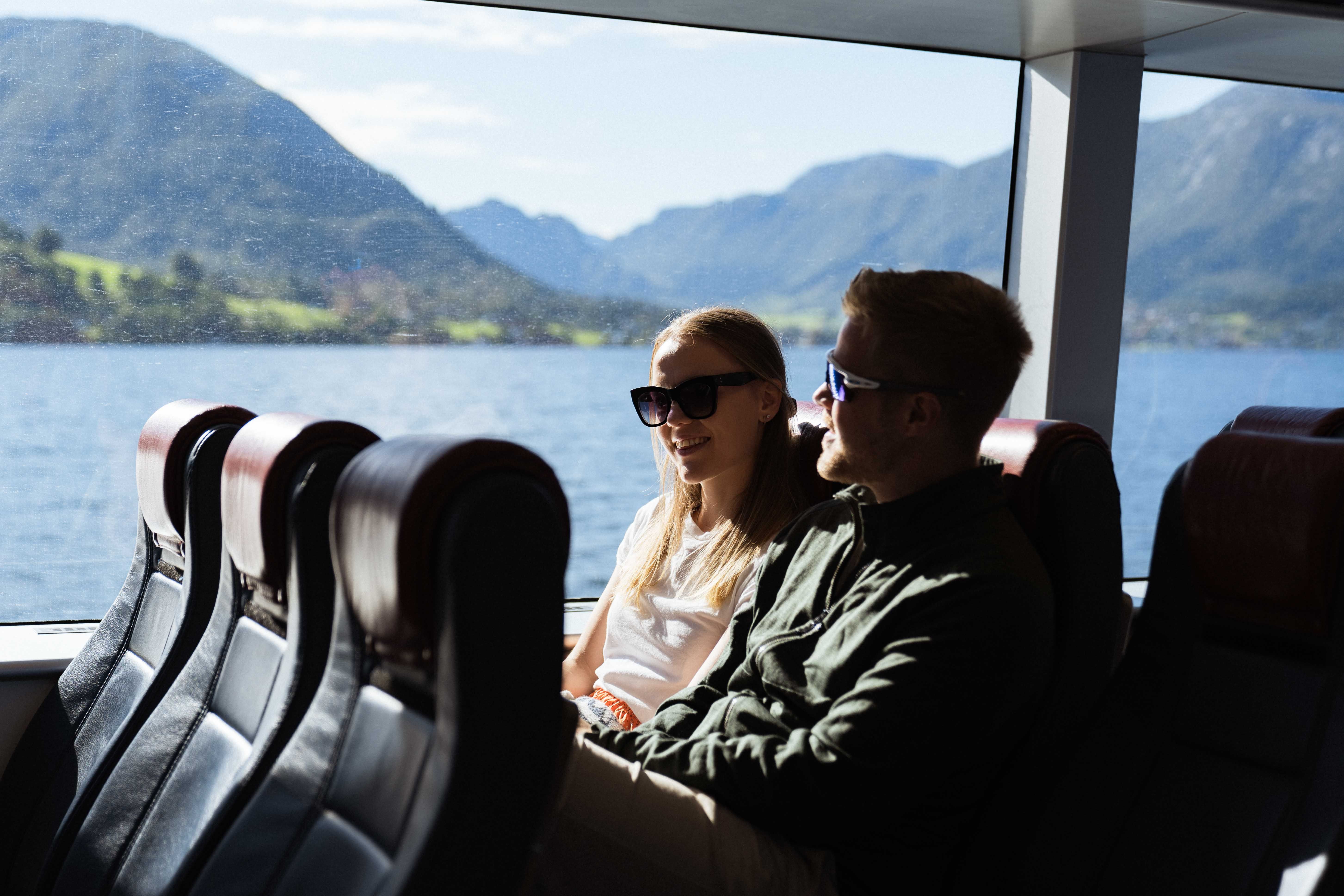 A couple sitting indoors on Hardangerfjordekspressen enjoying the fjord view through large windows.