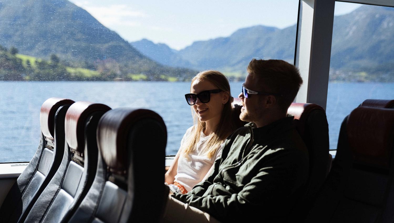 A couple sitting indoors on Hardangerfjordekspressen enjoying the fjord view through large windows.