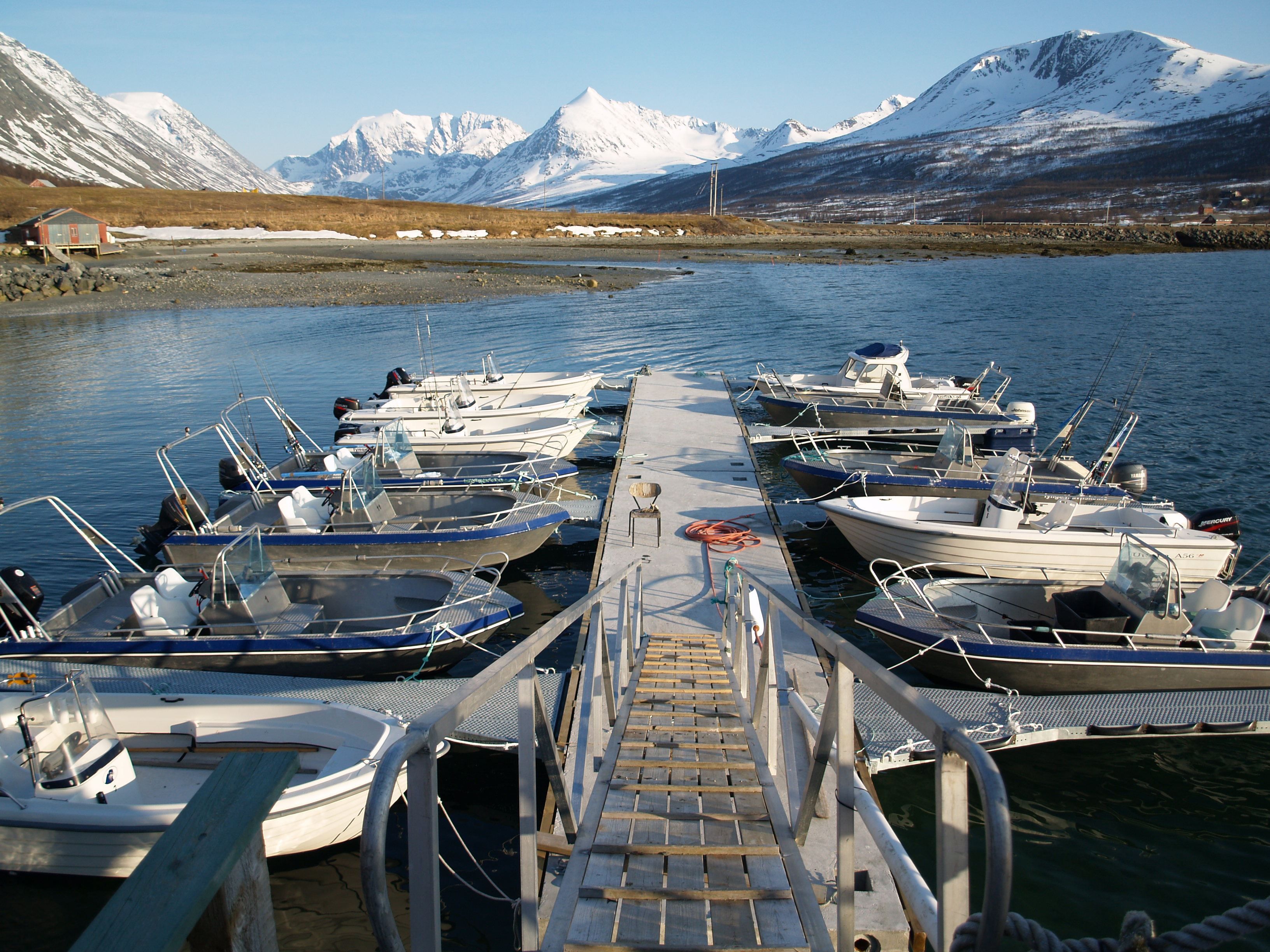 Jetty with small boats, a ramp, and a view of snow-covered mountains under a blue sky