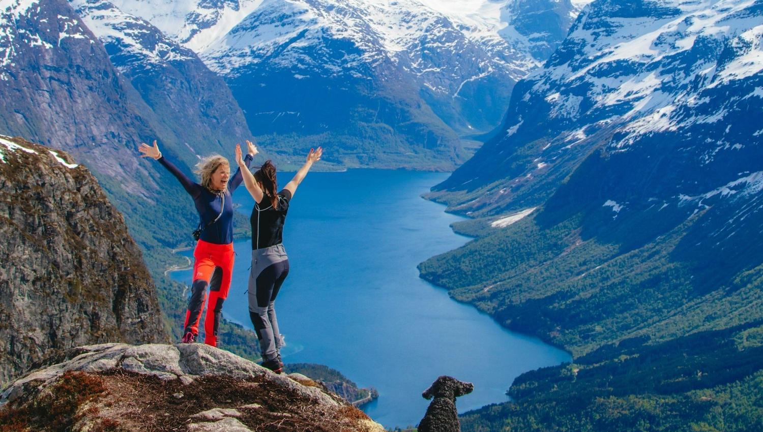Two hikers with raised arms stand on a rocky cliff overlooking a long blue lake and snowy mountains; a dog sits nearby.