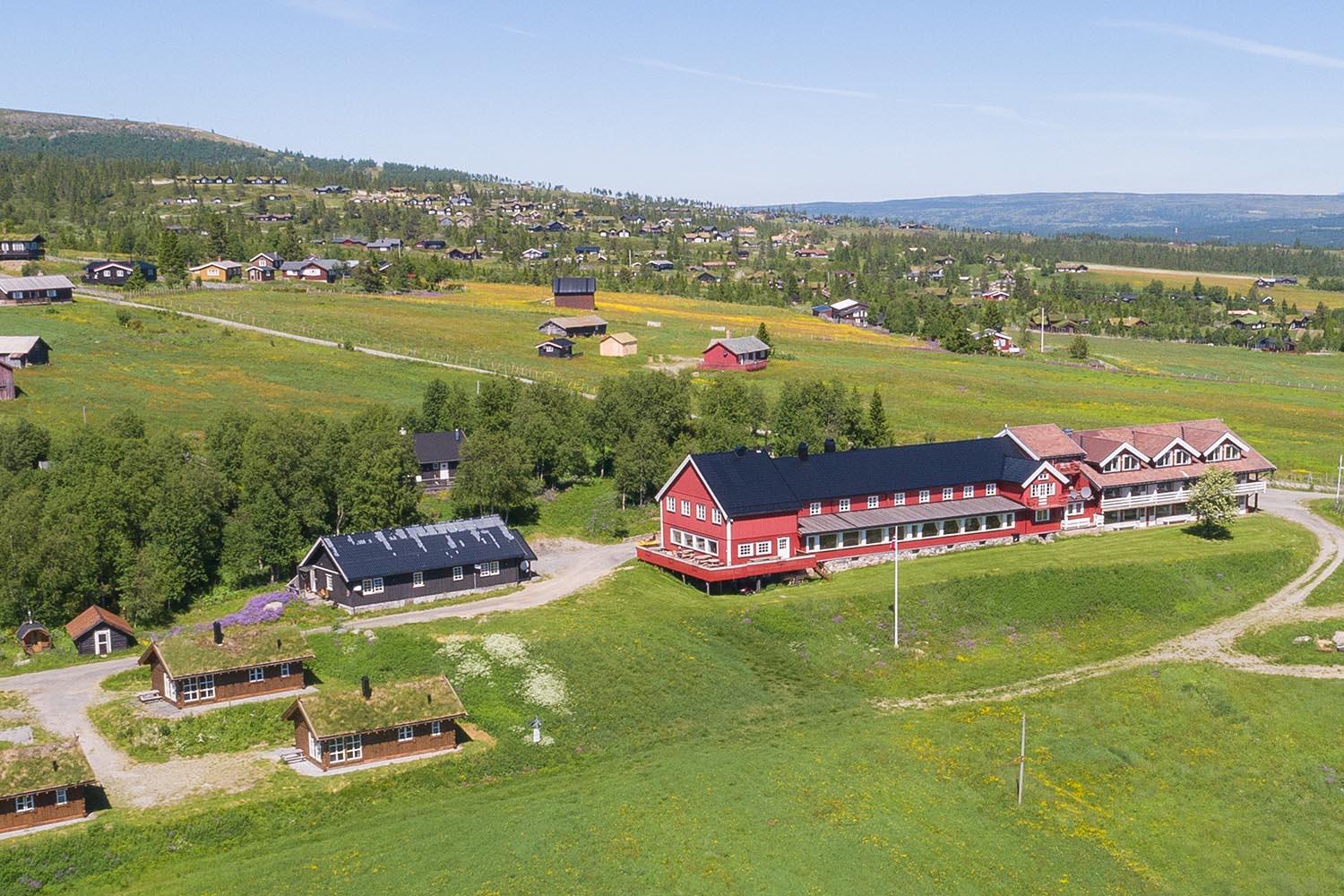Dronebilde av et rødt trehotell med noen hytter i åpent sommerlandskap med noen hytter og skog i bakgrunnen