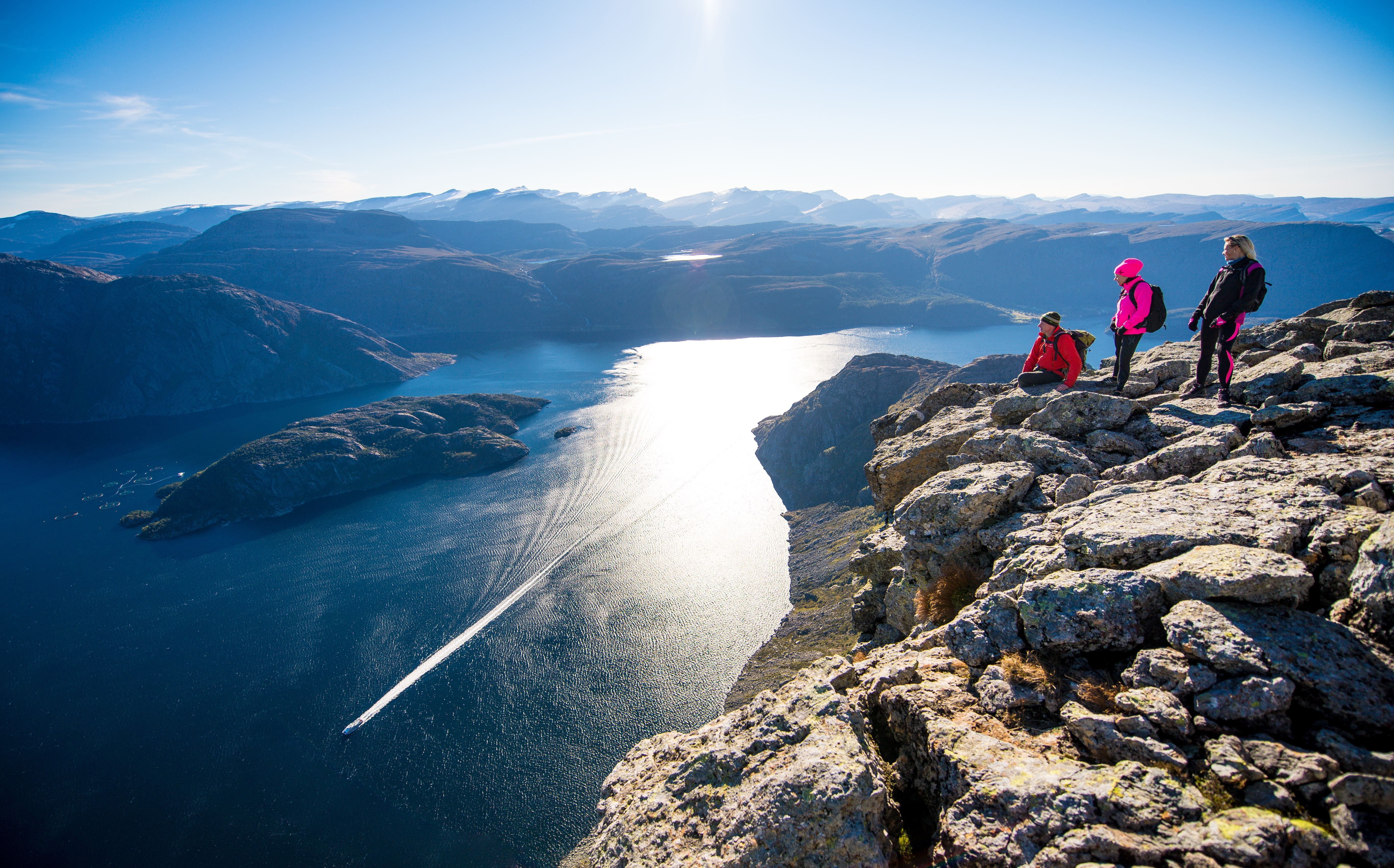 Tre vandrarar med ryggsekk står og sit på ei steinete fjellhylle med utsikt over ein solglitrande fjord og fjell..