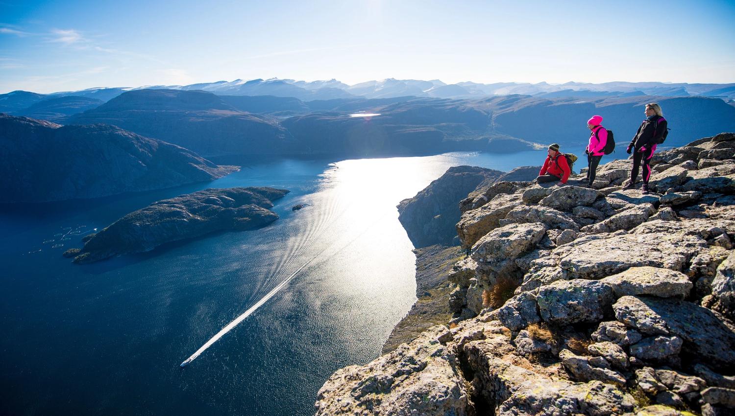 Tre vandrarar med ryggsekk står og sit på ei steinete fjellhylle med utsikt over ein solglitrande fjord og fjell..