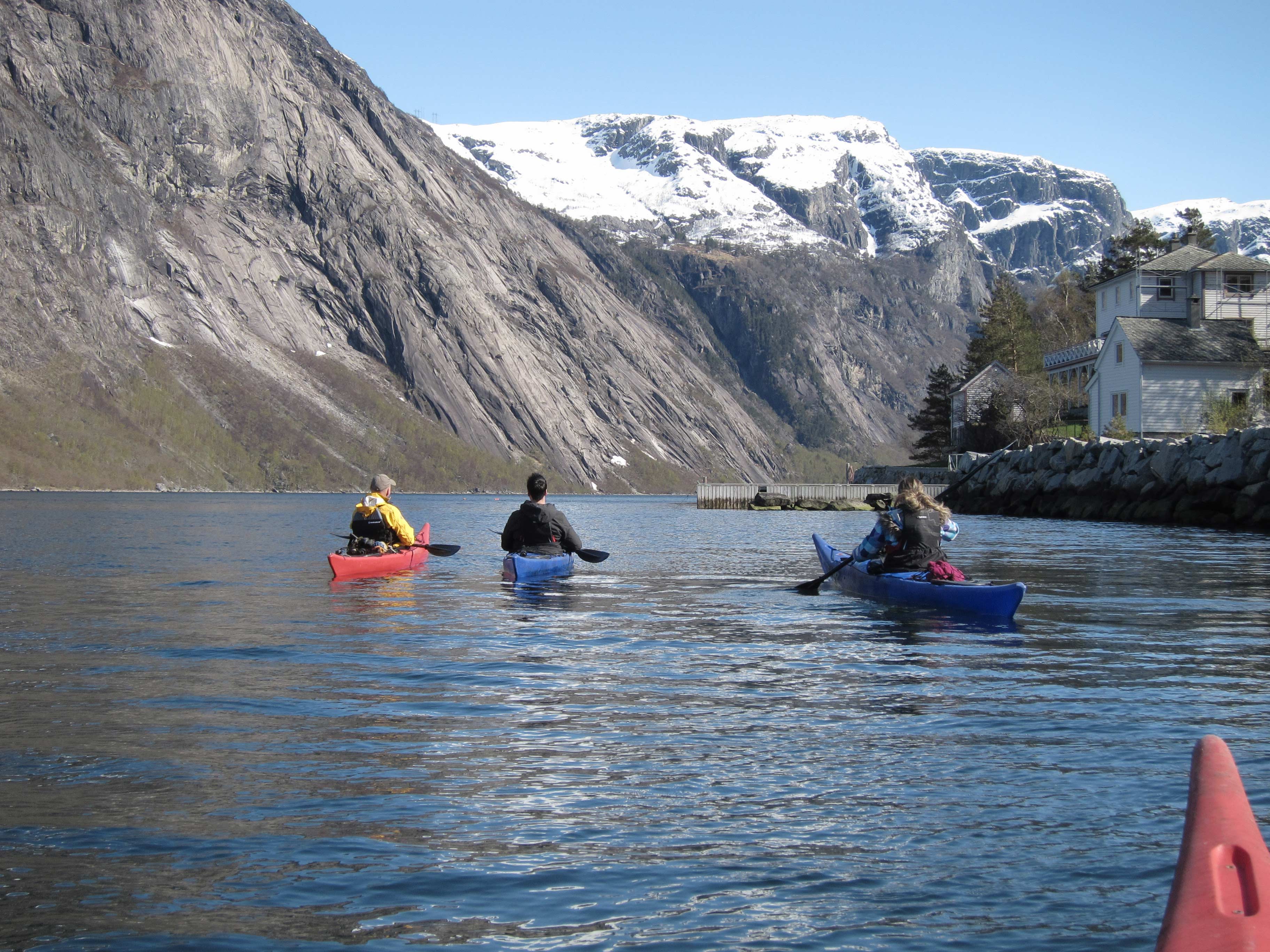 Kayakers in Eidfjord with snow-capped mountains in the background and a traditional Norwegian house by the fjord.