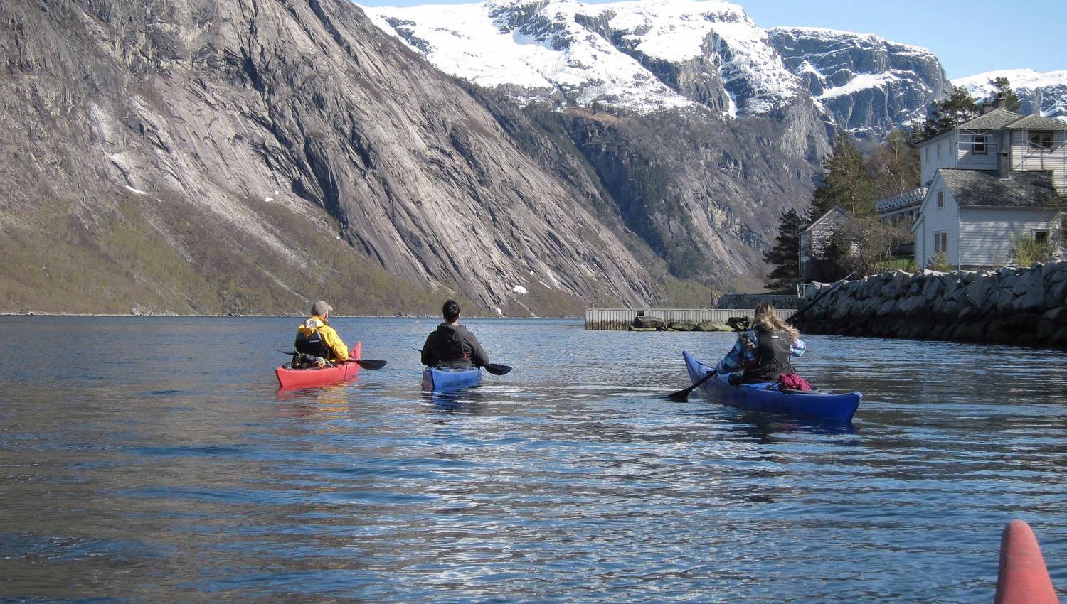 Kayakers in Eidfjord with snow-capped mountains in the background and a traditional Norwegian house by the fjord.