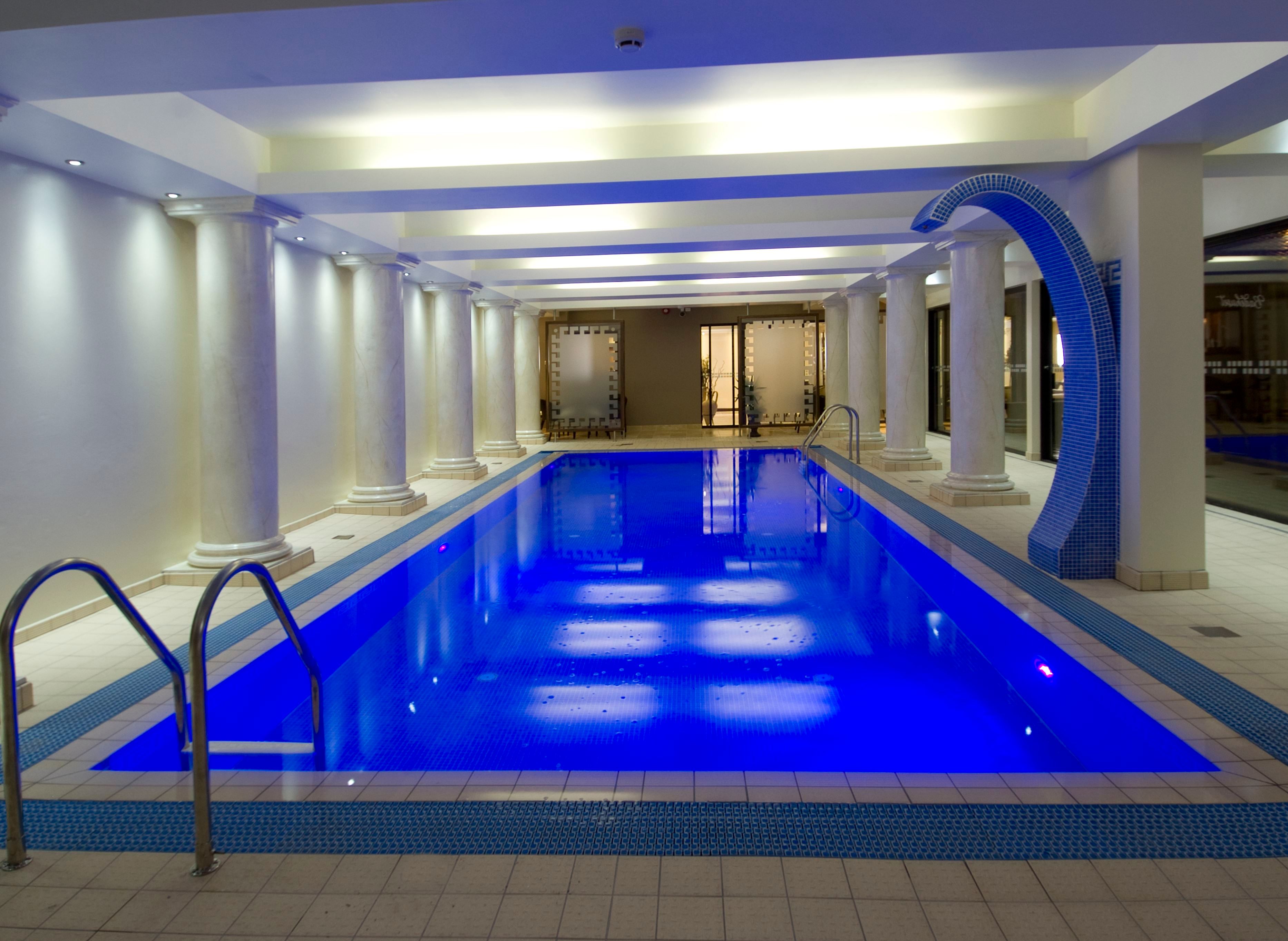 Long indoor swimming pool with blue lighting, tiled deck, columns along the sides, ladders, and a curved water feature.