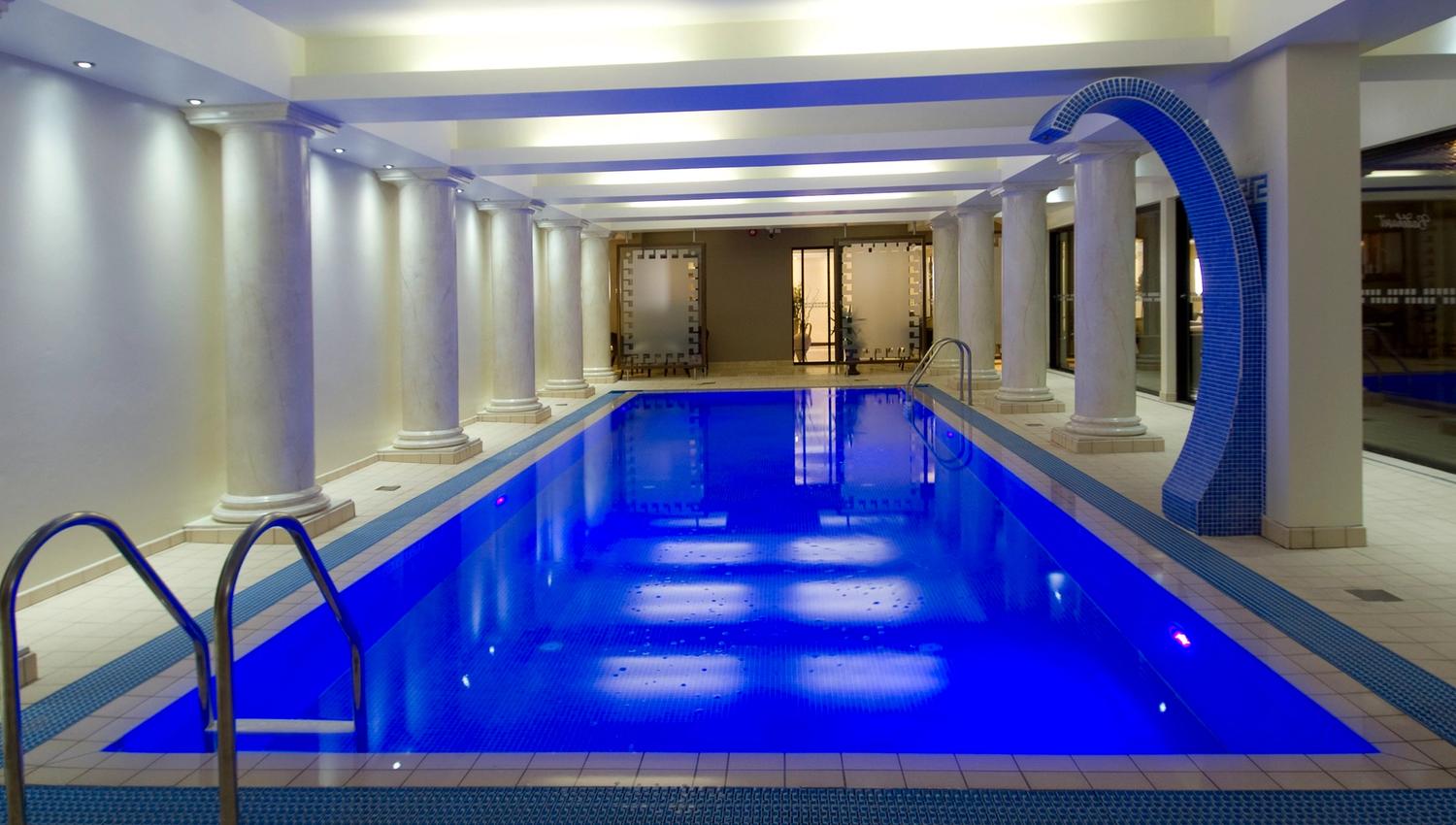 Long indoor swimming pool with blue lighting, tiled deck, columns along the sides, ladders, and a curved water feature.