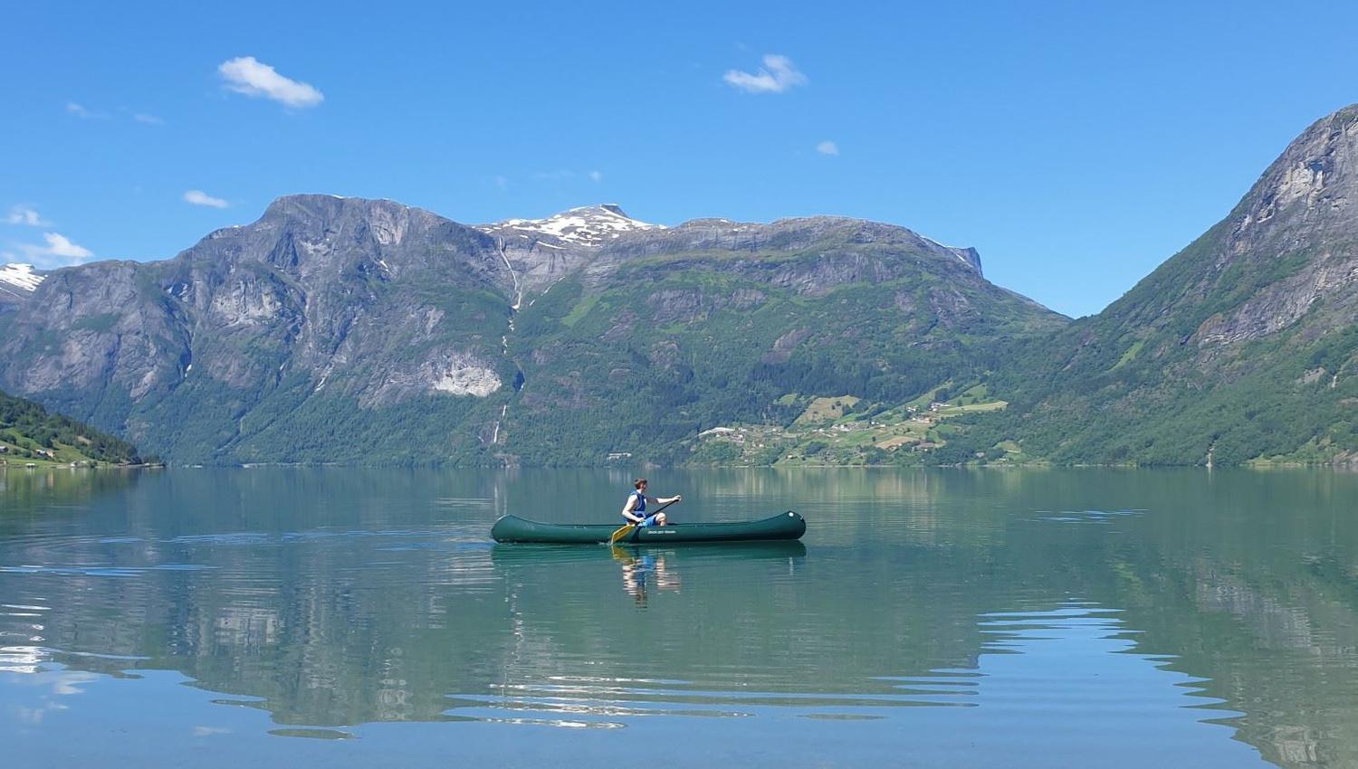 Ein person padlar ein grøn kano på spegelblankt vatn framfor grøne fjell med snøflekkar og blå himmel..