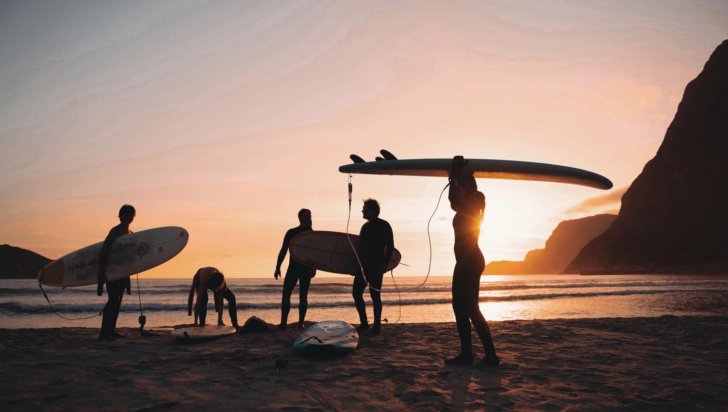 Silhouetted surfers with surfboards on a sandy beach at sunset, with ocean waves and coastal cliffs in the background by Hoddevik Surf House at Stadlandet..