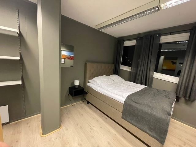 Minimalist bedroom with a single bed, gray walls, built-in shelves, nightstand lamp, and curtained windows.