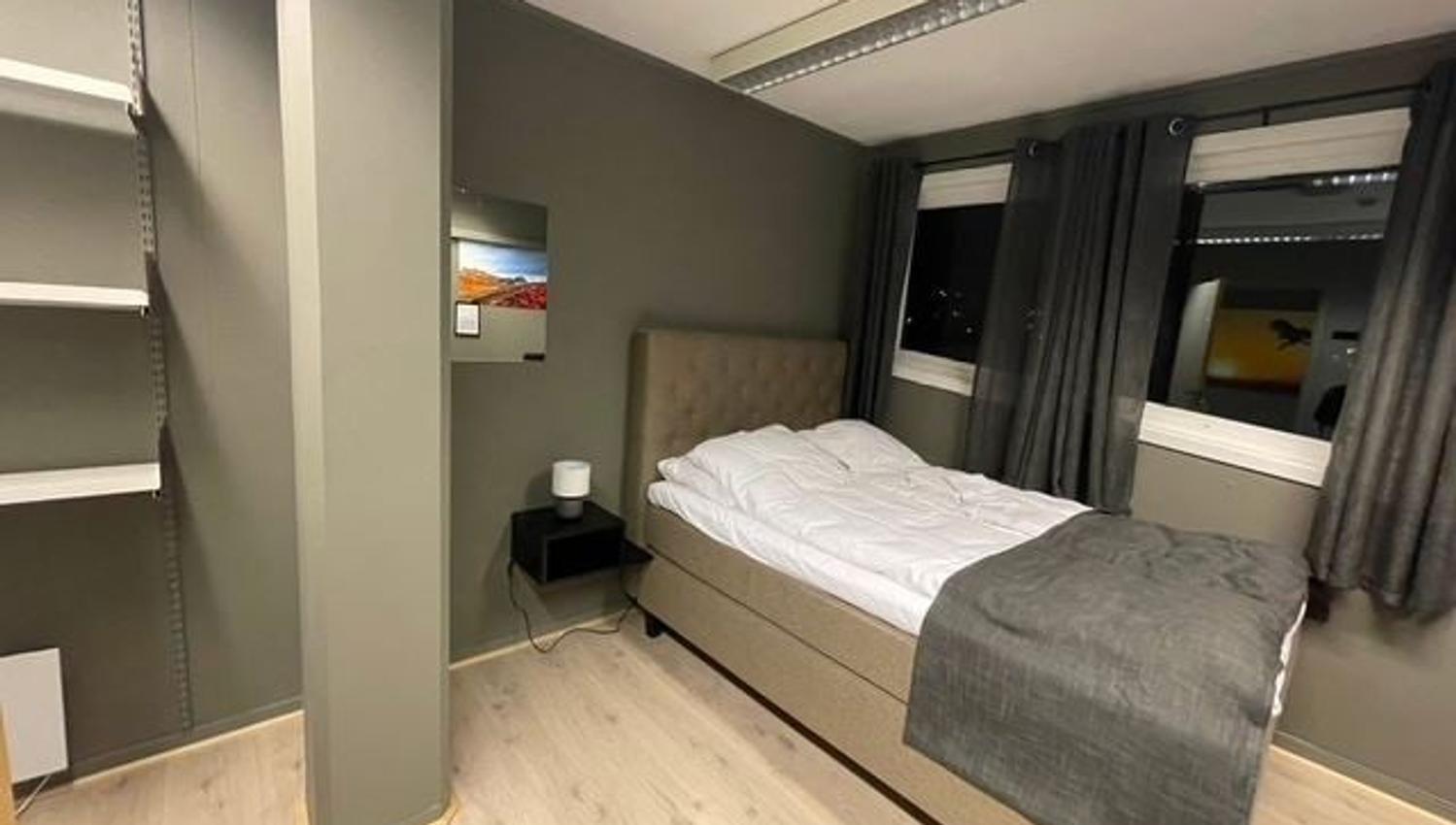 Minimalist bedroom with a single bed, gray walls, built-in shelves, nightstand lamp, and curtained windows.