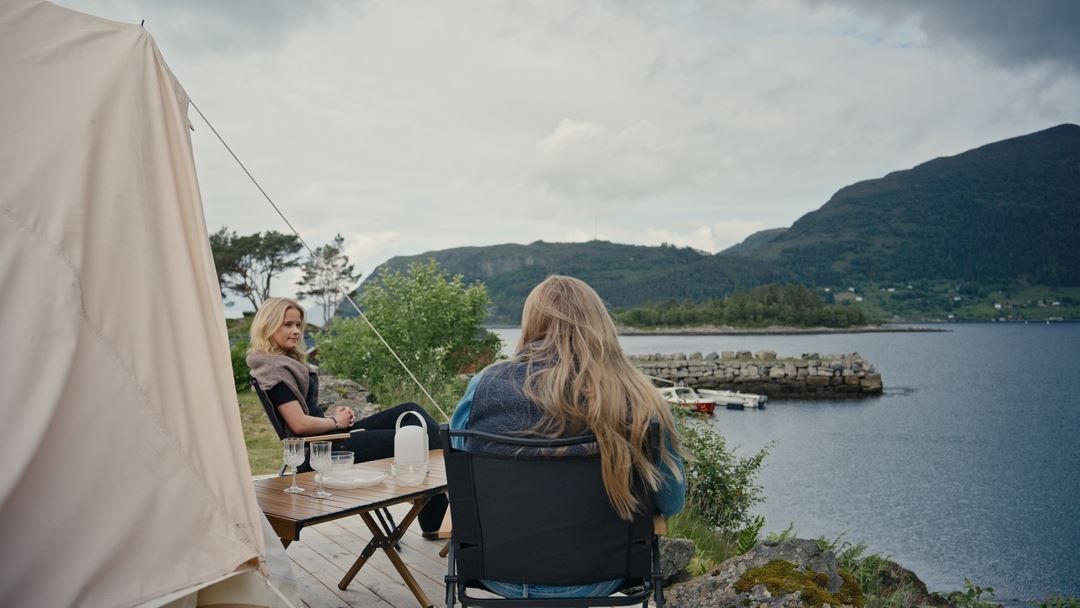 Two women sit by a canvas tent on a wooden deck overlooking a calm lake, rocky shore, and distant mountains..