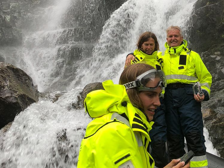 Excited participants stand close to the waterfall, feeling the powerful spray on a RIB safari in Hardanger.