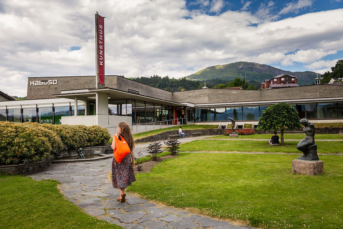 Kunsthuset Kabuso in Hardanger, with a well-kept sculpture park, a woman with an orange bag in the foreground, and mountains in the background.