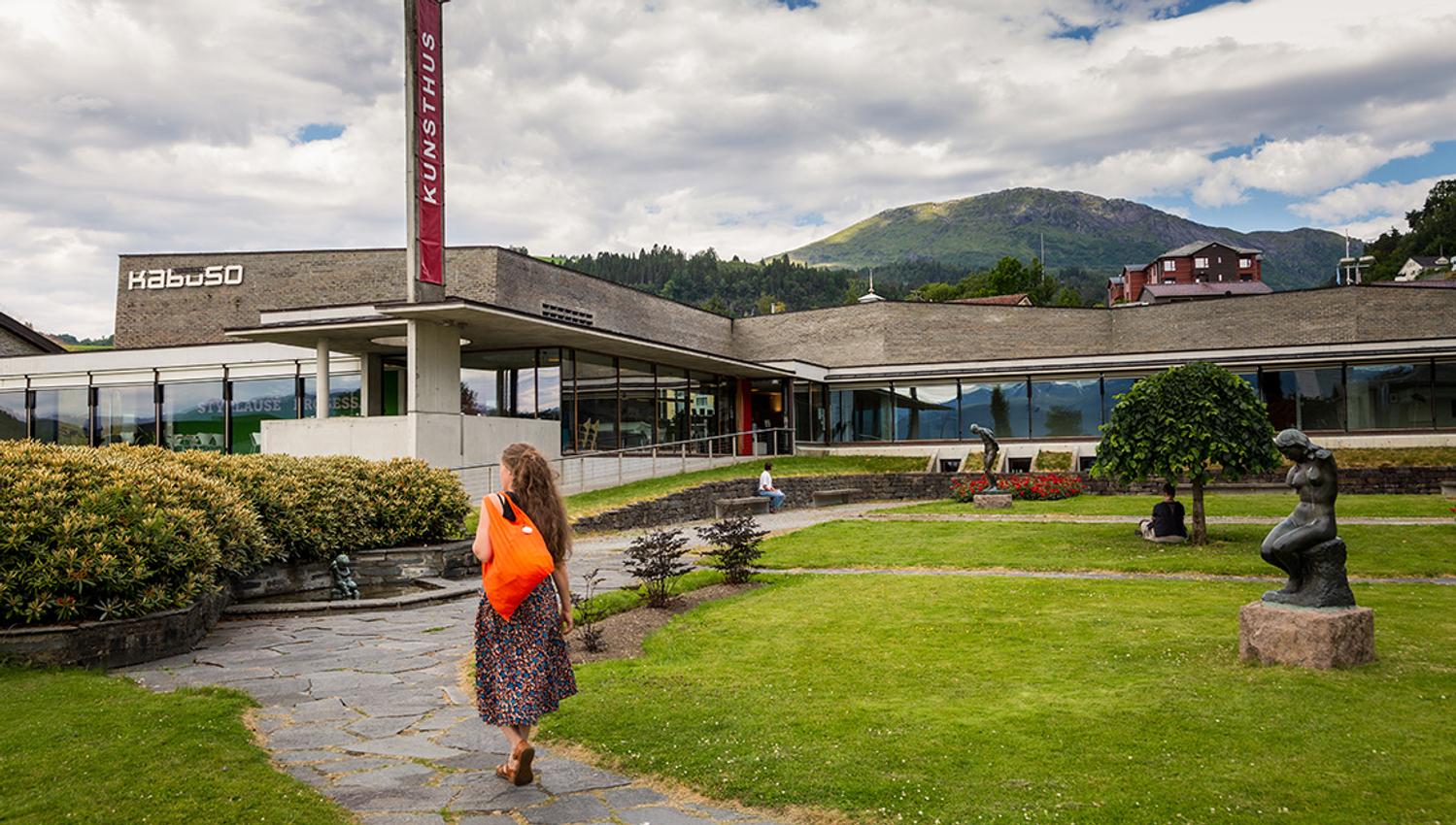 Kunsthuset Kabuso in Hardanger, with a well-kept sculpture park, a woman with an orange bag in the foreground, and mountains in the background.