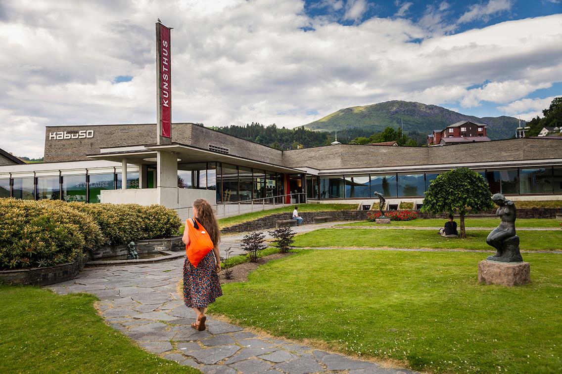 Kunsthuset Kabuso in Hardanger, with a well-kept sculpture park, a woman with an orange bag in the foreground, and mountains in the background.