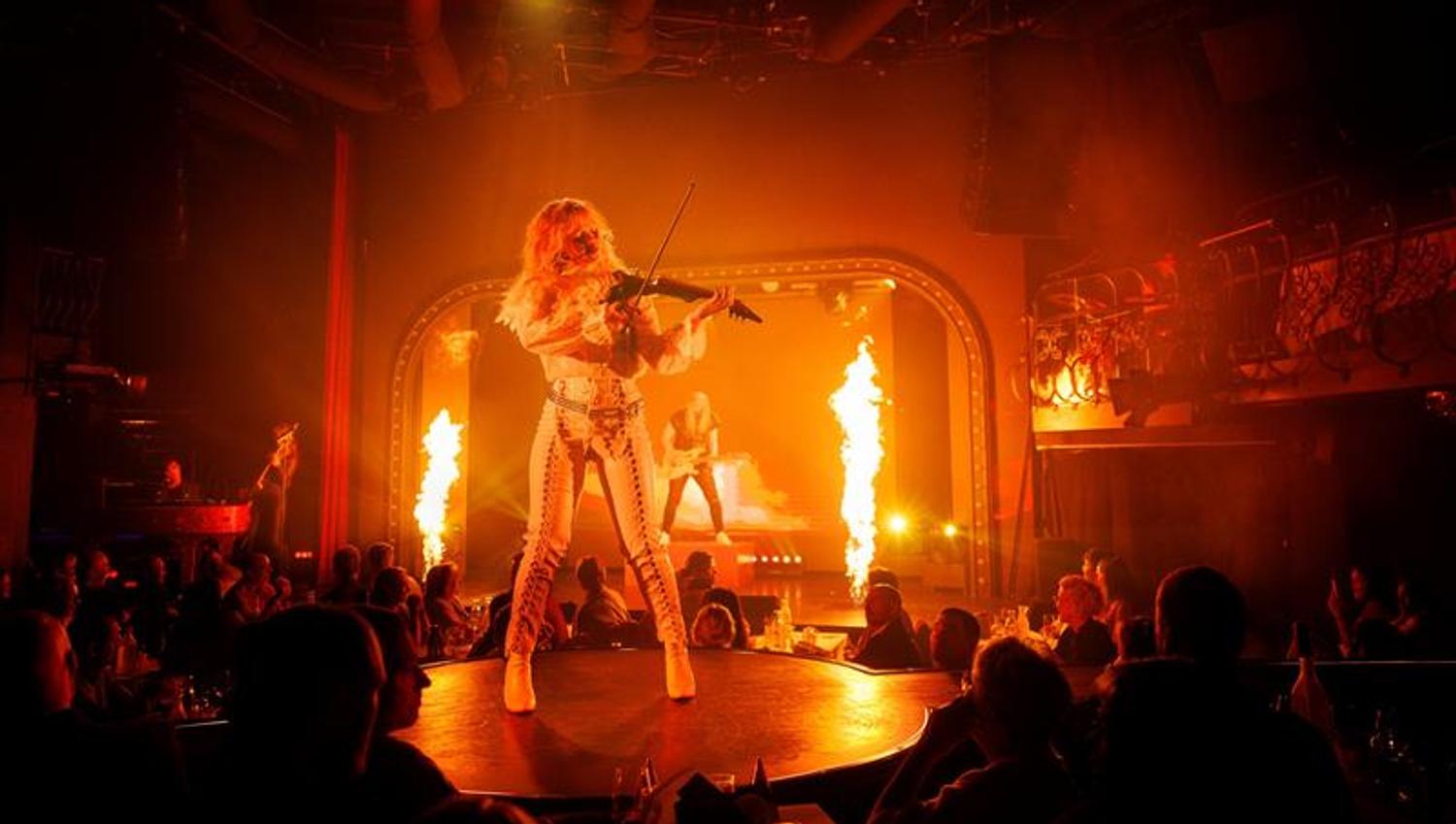 En fiolinist opptrer på en rund scene omgitt av flammer og publikum, mens en gitarist spiller i bakgrunnen. Scenen gløder i varmt oransje lys.