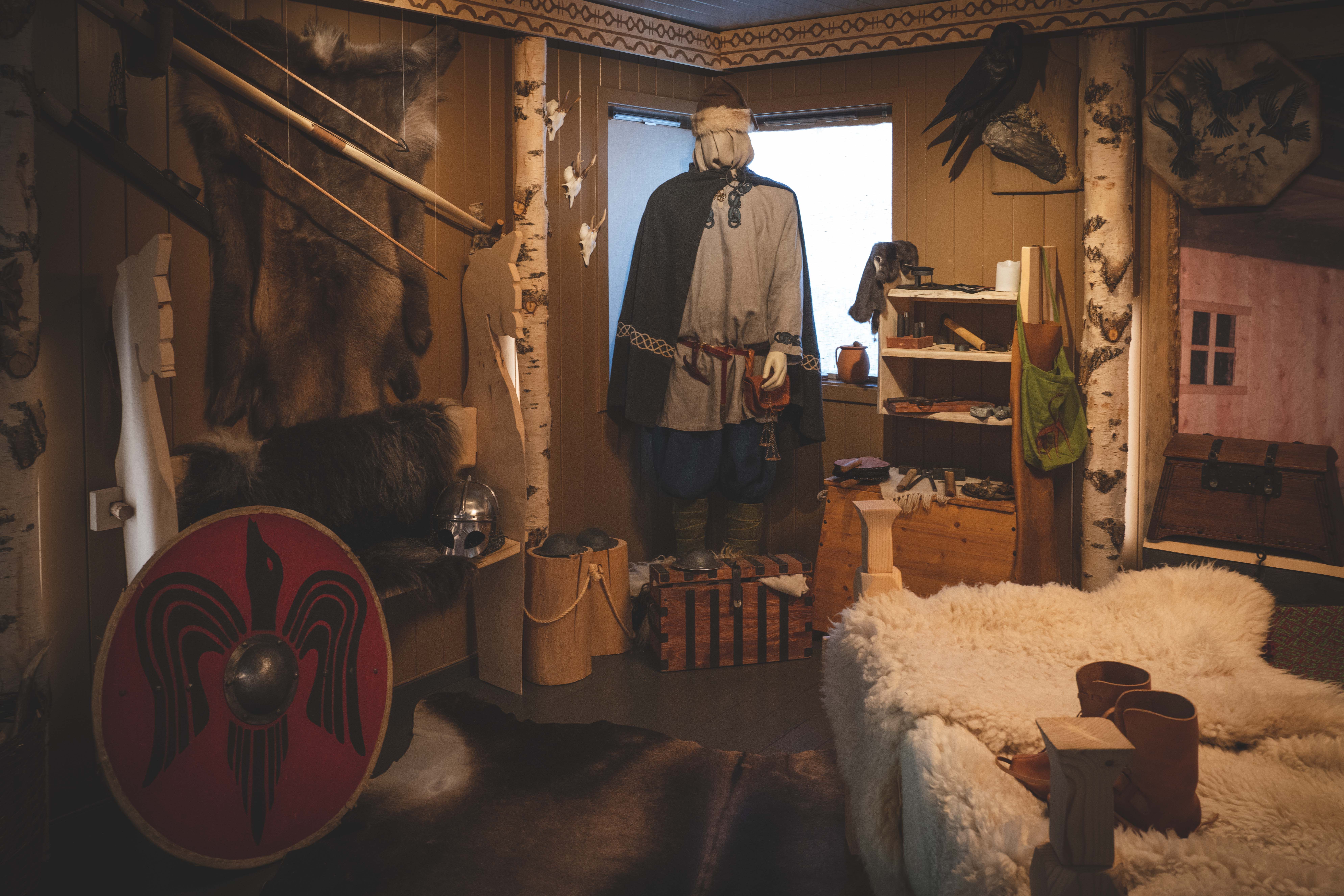 In der Wikingerausstellung hängen ein Schild, eine Schaufensterpuppe in Wikingerkleidung, ein Bett mit Schaffellen und ein Rentierfell an der Wand.