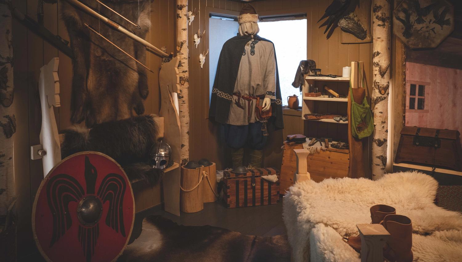 In der Wikingerausstellung hängen ein Schild, eine Schaufensterpuppe in Wikingerkleidung, ein Bett mit Schaffellen und ein Rentierfell an der Wand.