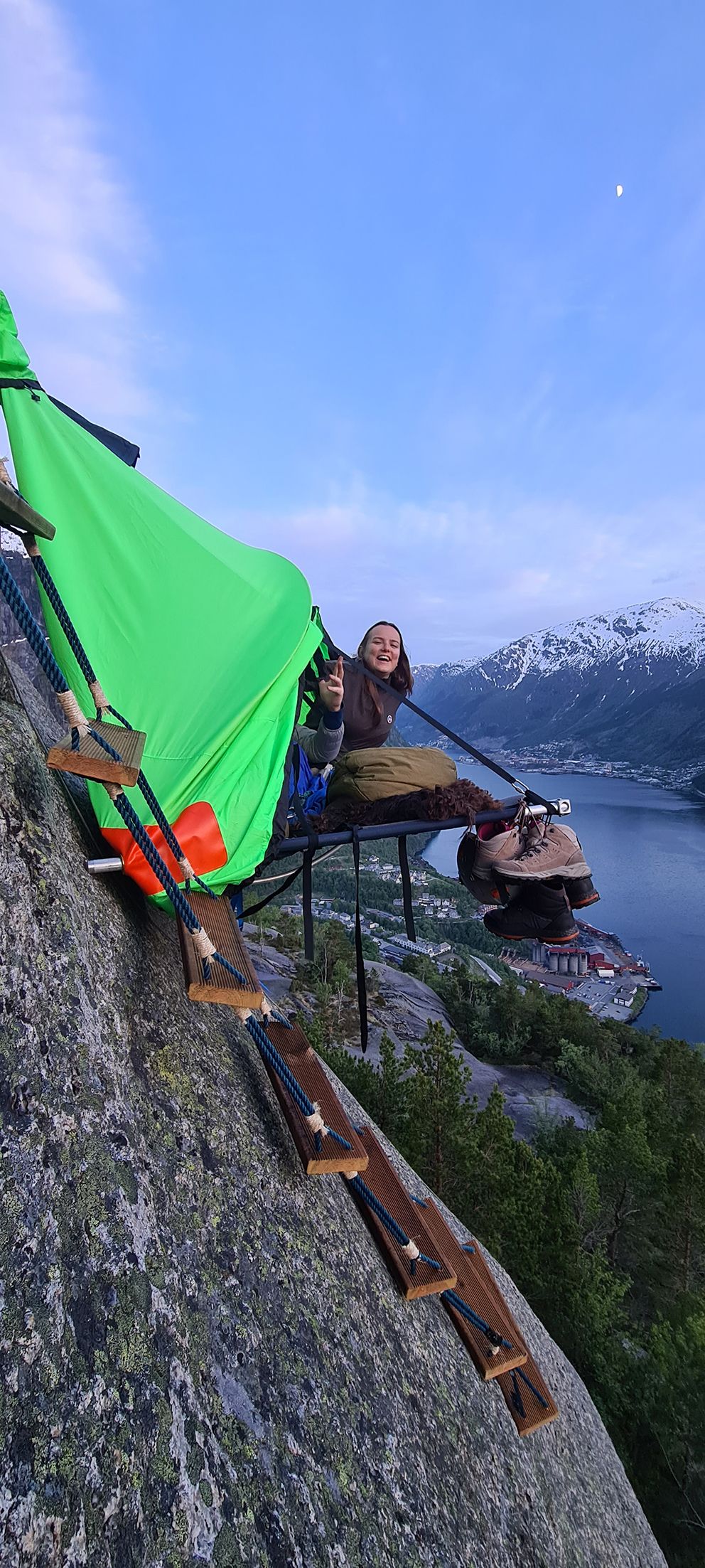 Ei kvinne smiler frå ein portaledge med spektakulær utsikt over fjorden og fjella i Hardanger. Eit adrenalinfylt overnattingseventyr.