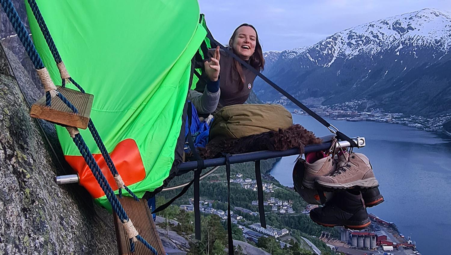 Ei kvinne smiler frå ein portaledge med spektakulær utsikt over fjorden og fjella i Hardanger. Eit adrenalinfylt overnattingseventyr.