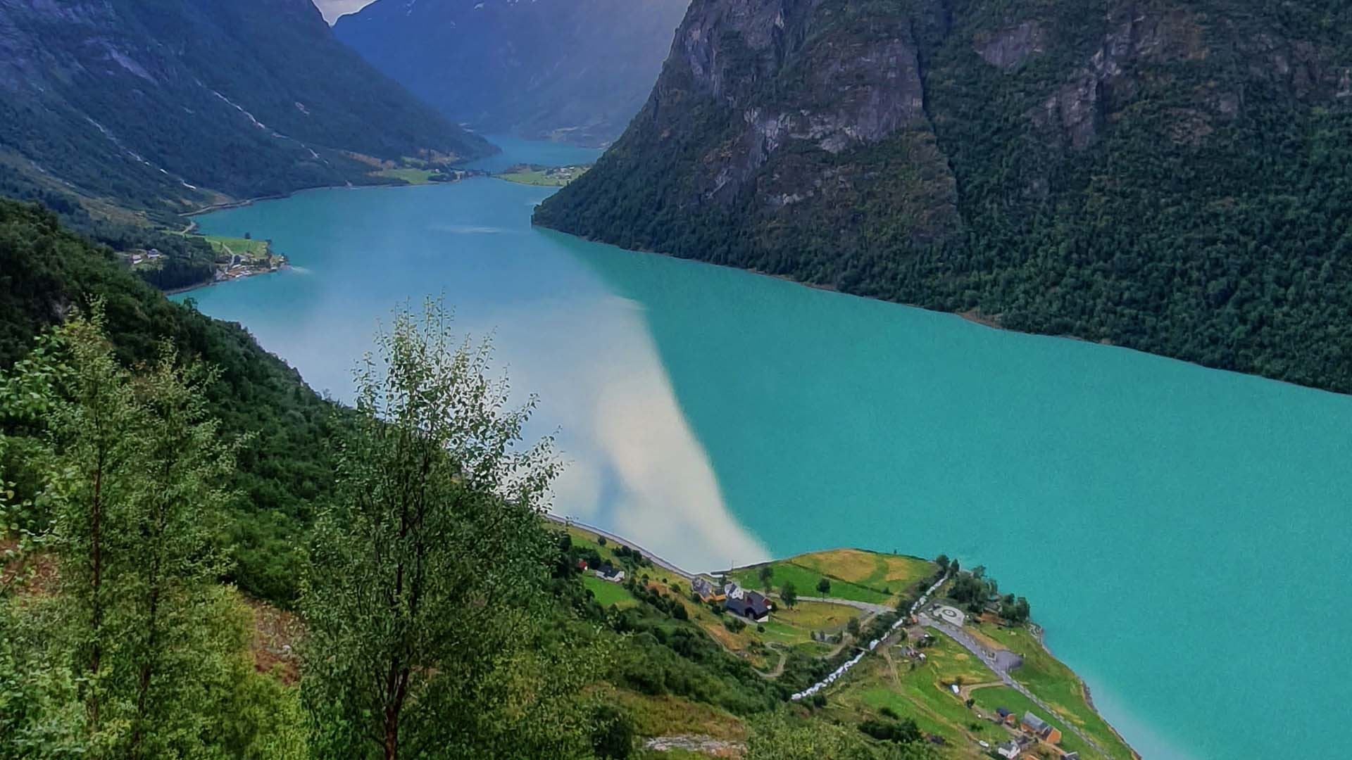 Elevated view of a turquoise fjord flanked by steep green mountains, with a small village and roads on shore.