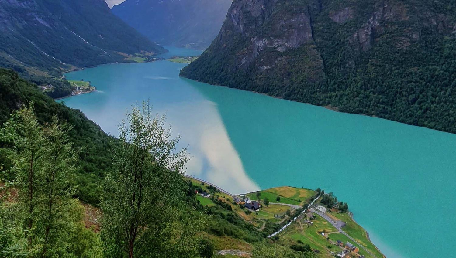 Elevated view of a turquoise fjord flanked by steep green mountains, with a small village and roads on shore.