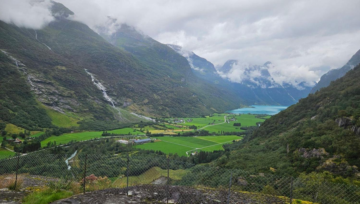 Wide view of a green valley between mountains, with waterfalls, farmland, and a turquoise fjord under clouds..