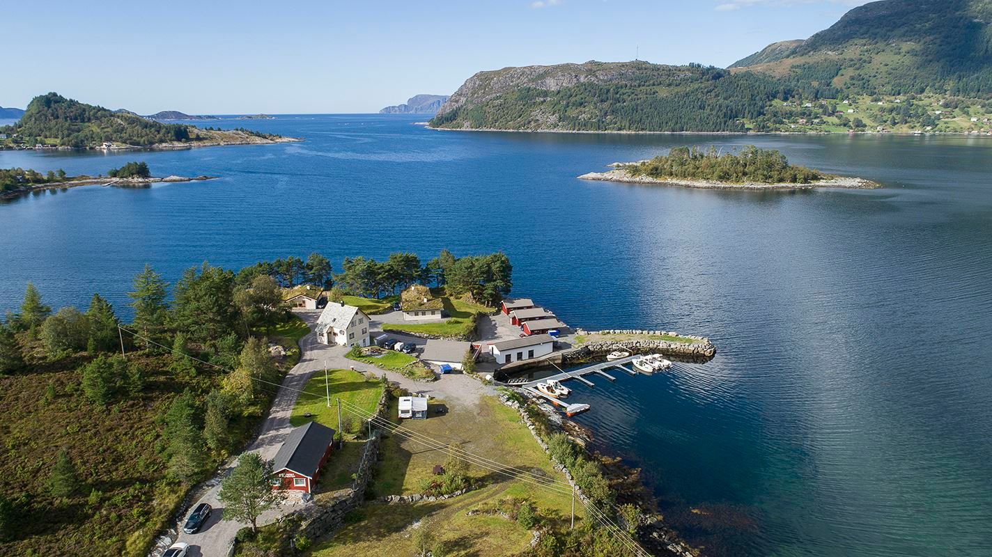 Aerial view of a small coastal settlement with houses and a dock with boats beside calm blue water and green mountains..