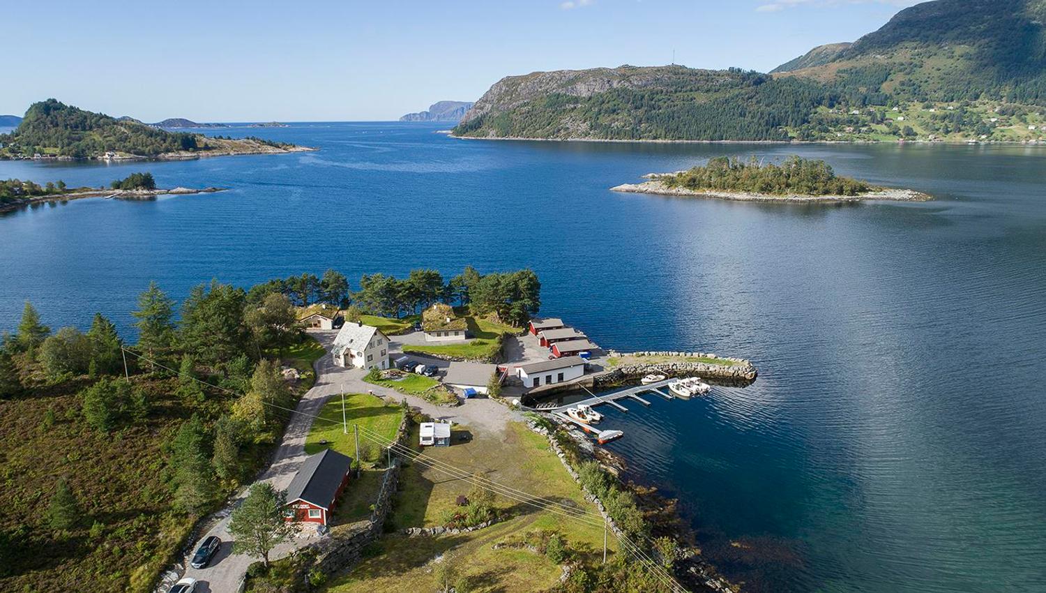 Aerial view of a small coastal settlement with houses and a dock with boats beside calm blue water and green mountains..