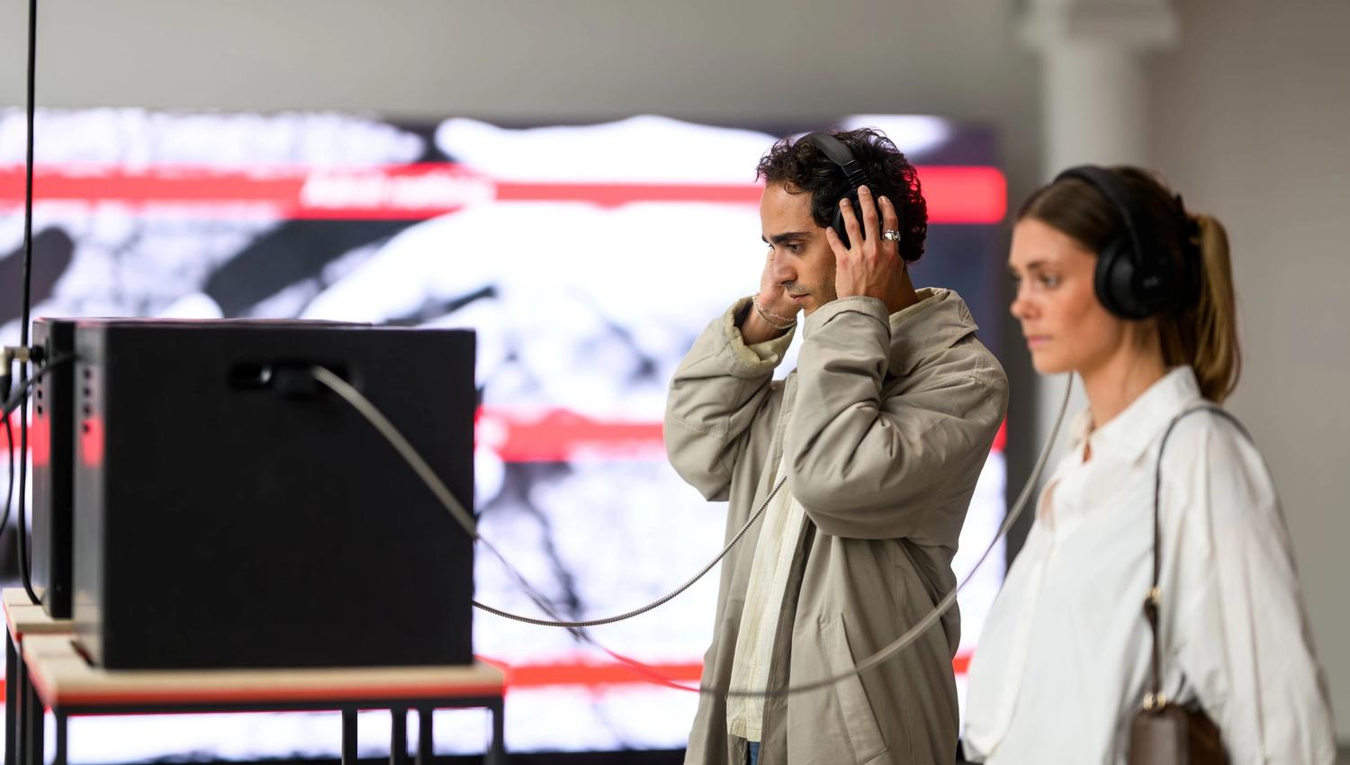 Two people wearing headphones experience a sound installation at the Nobel Peace Center. A man in a light coat stands focused in front of the equipmen