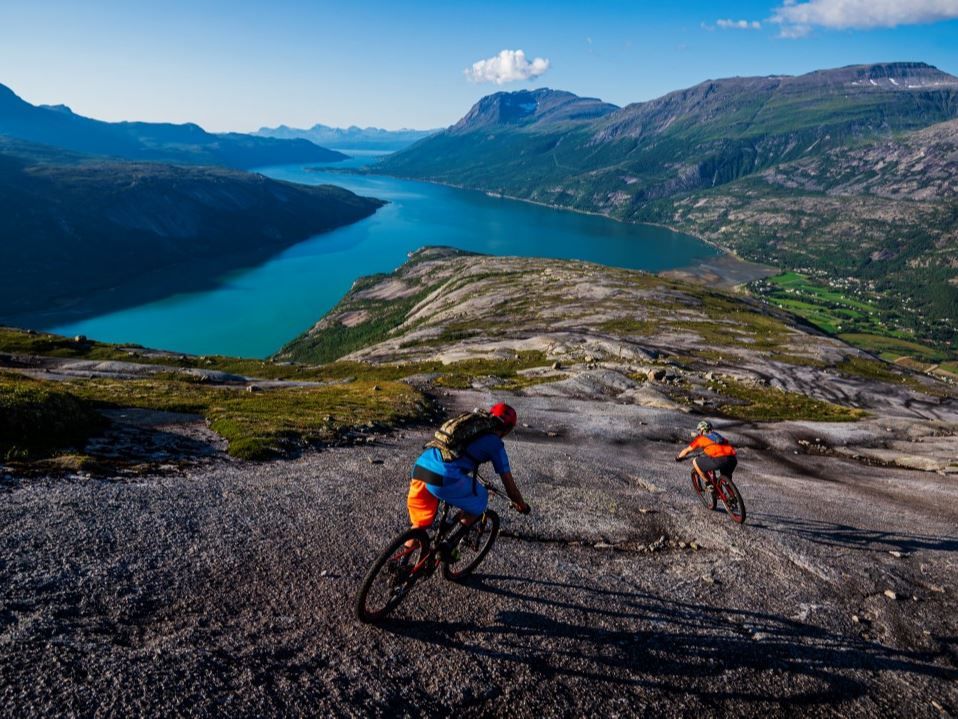 Two mountain bikers cycle down a rocky mountain plateau with turquoise fjords and mountains in the background.