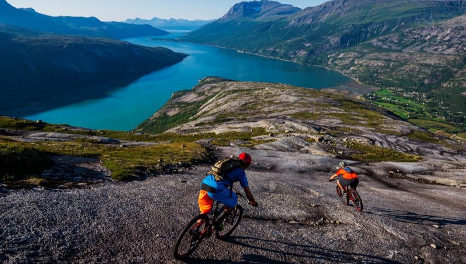 Two mountain bikers cycle down a rocky mountain plateau with turquoise fjords and mountains in the background.