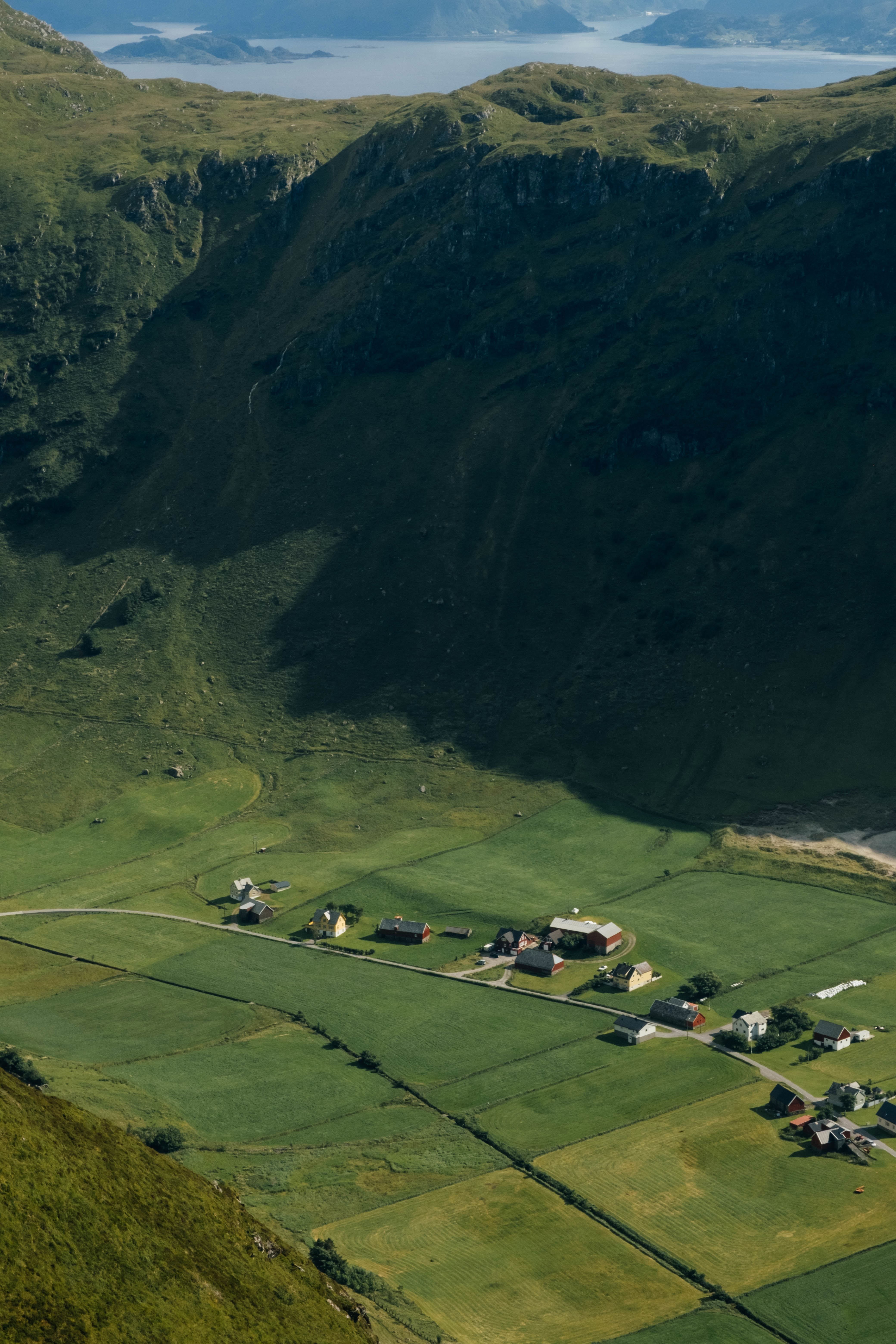 Aerial view of green fields and small farmhouses in a valley, shaded by steep mountains with water in the distance.