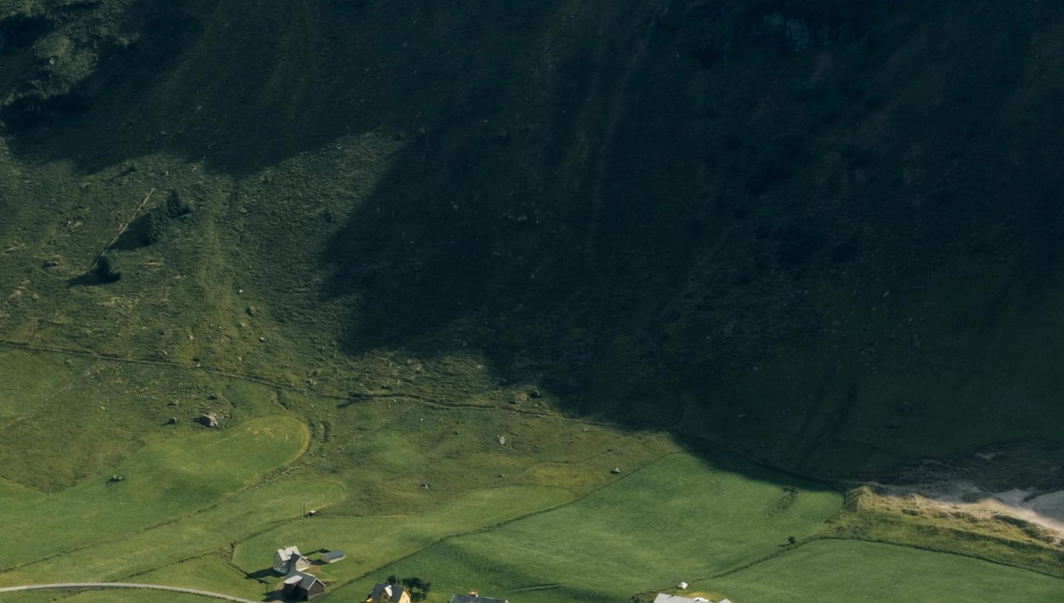 Aerial view of green fields and small farmhouses in a valley, shaded by steep mountains with water in the distance.