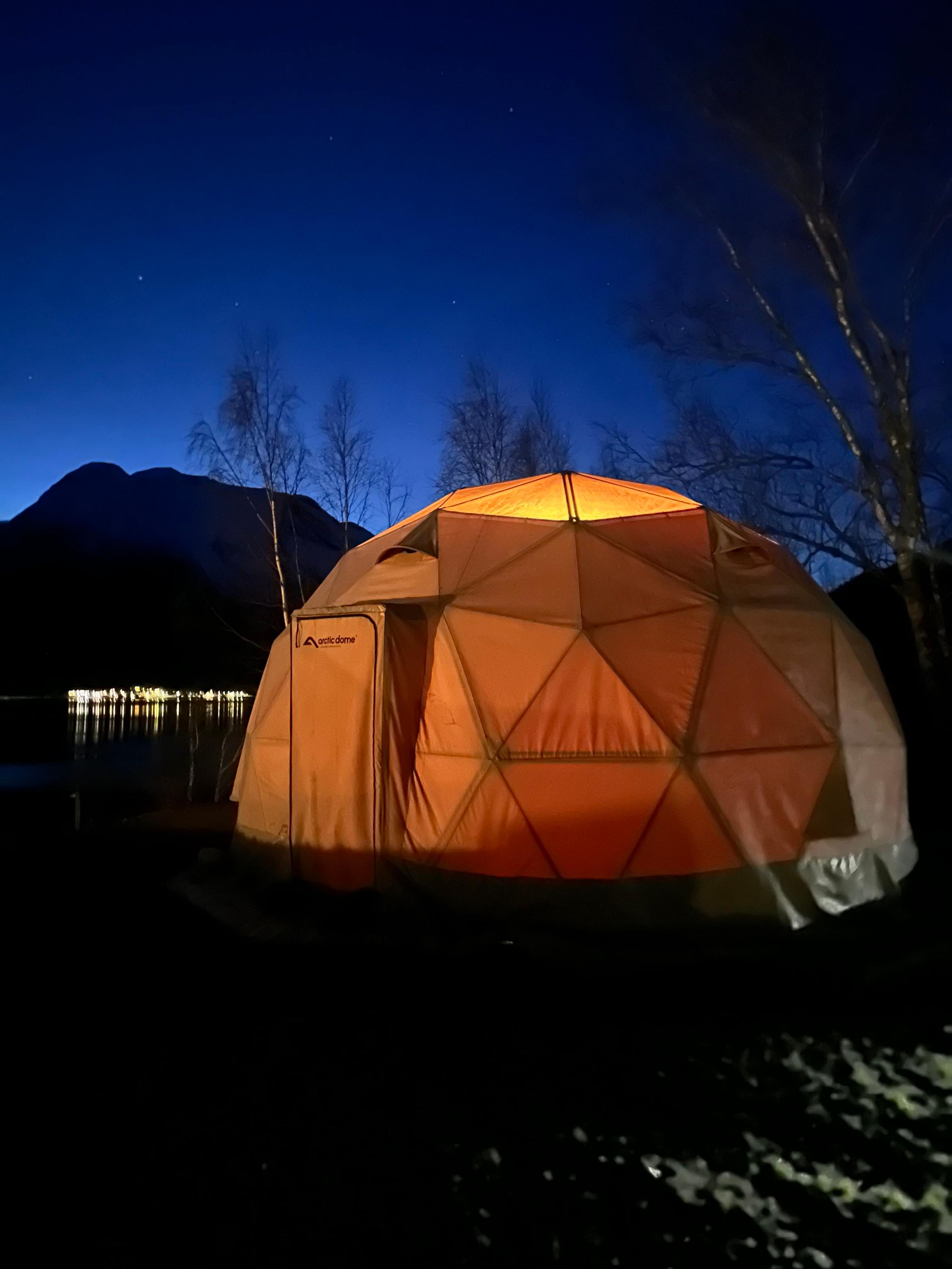 Eit opplyst oransje kuppeltelt står ved ei mørk strand, med fjell, nakne tre og stjernehimmel bak på Vollsnes i Oppstryn ved Oppstrynsvatnet..