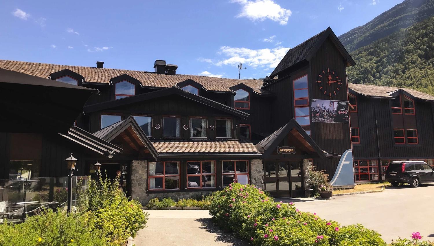 Dark wooden lodge with red-trim windows and a clock tower, front walkway and flower beds under a blue sky..