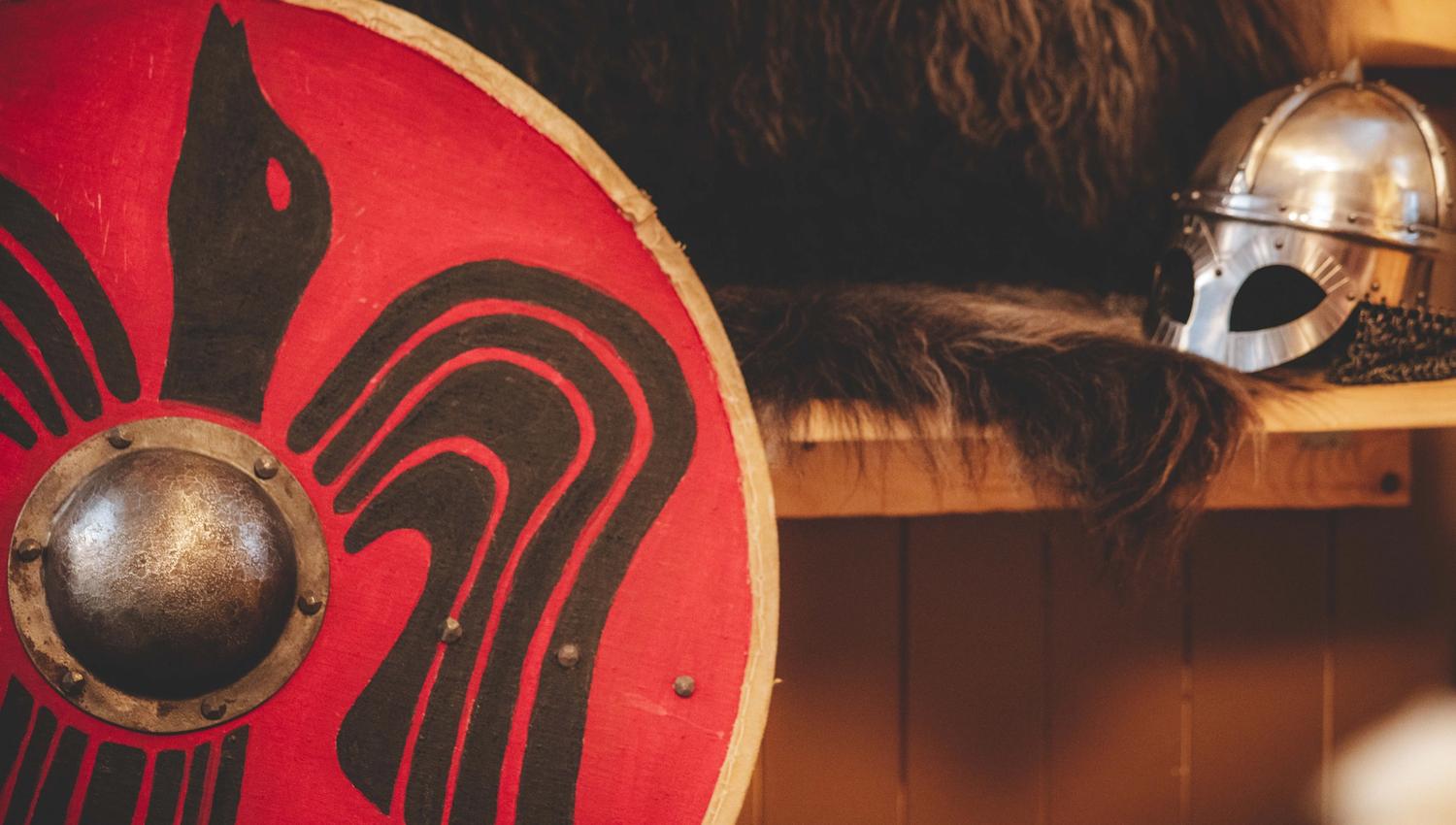 Eine Nahaufnahme eines Wikingerschildes, im Hintergrund ist ein Wikingerhelm zu sehen.