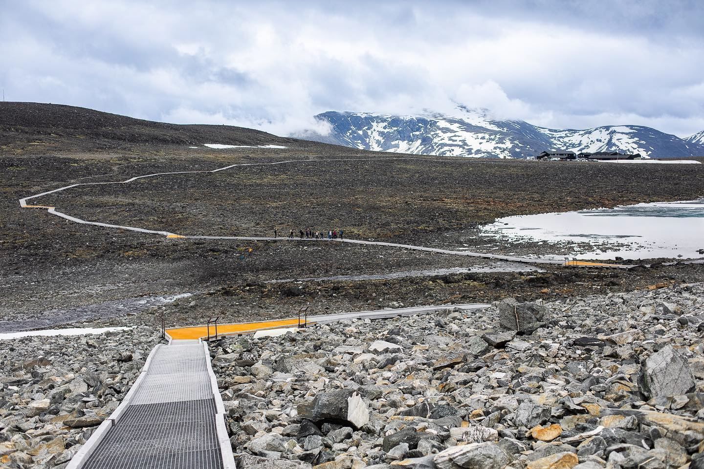Lang plankesti kryssar steinete fjellvidde mot snødekte fjell, med lite vatn til høgre og skyer over..