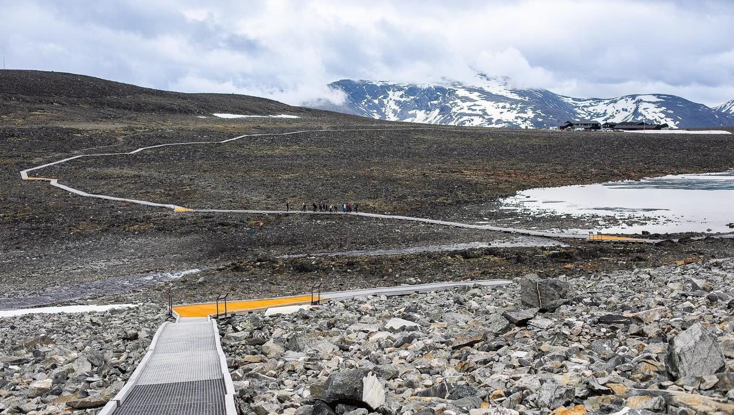 Lang plankesti kryssar steinete fjellvidde mot snødekte fjell, med lite vatn til høgre og skyer over..