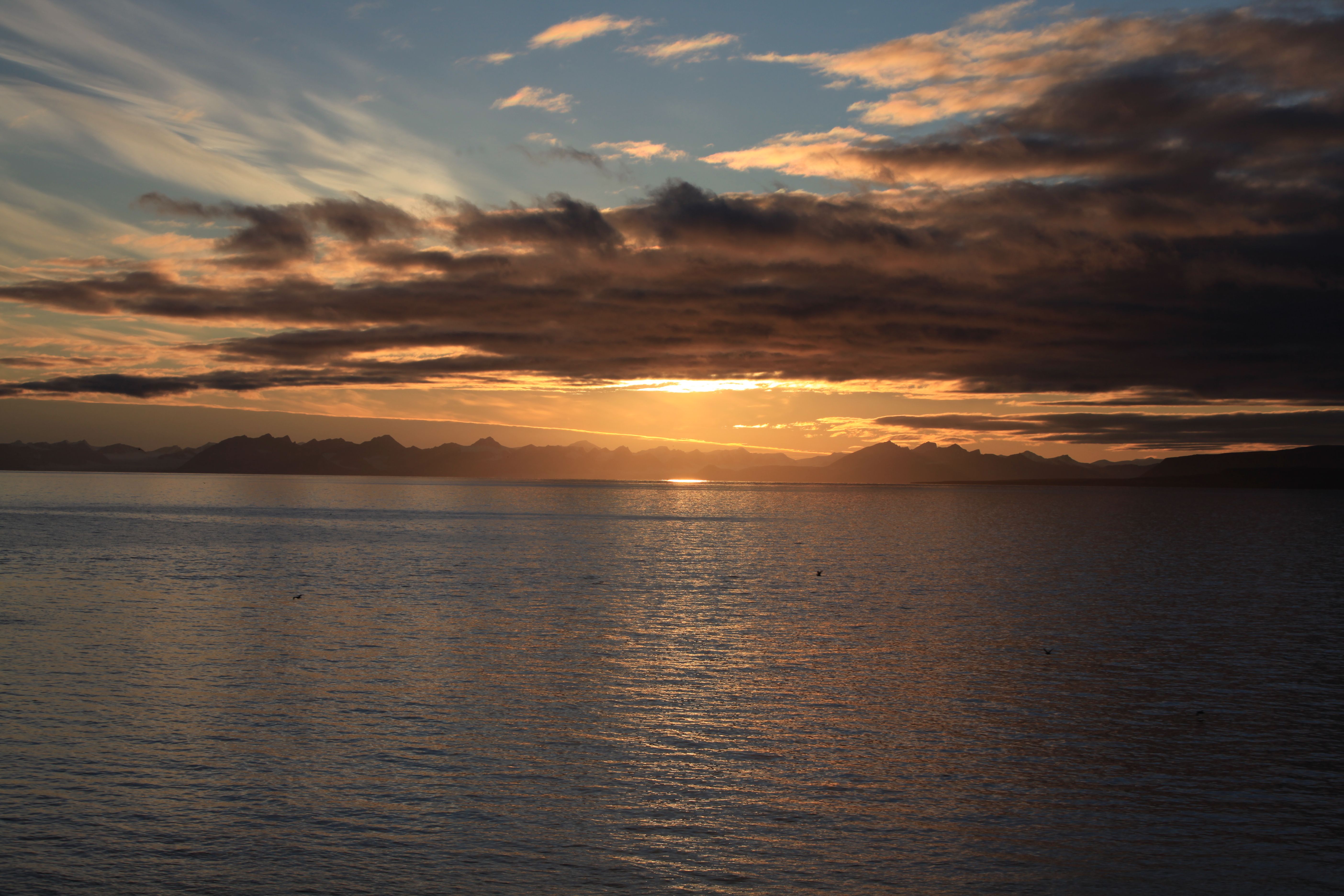 A dark fjord and dark skies with a setting sun and mountains in silhouette in the background