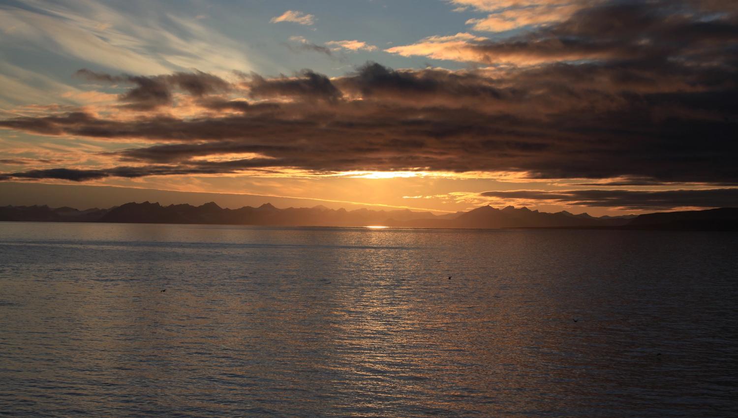 A dark fjord and dark skies with a setting sun and mountains in silhouette in the background