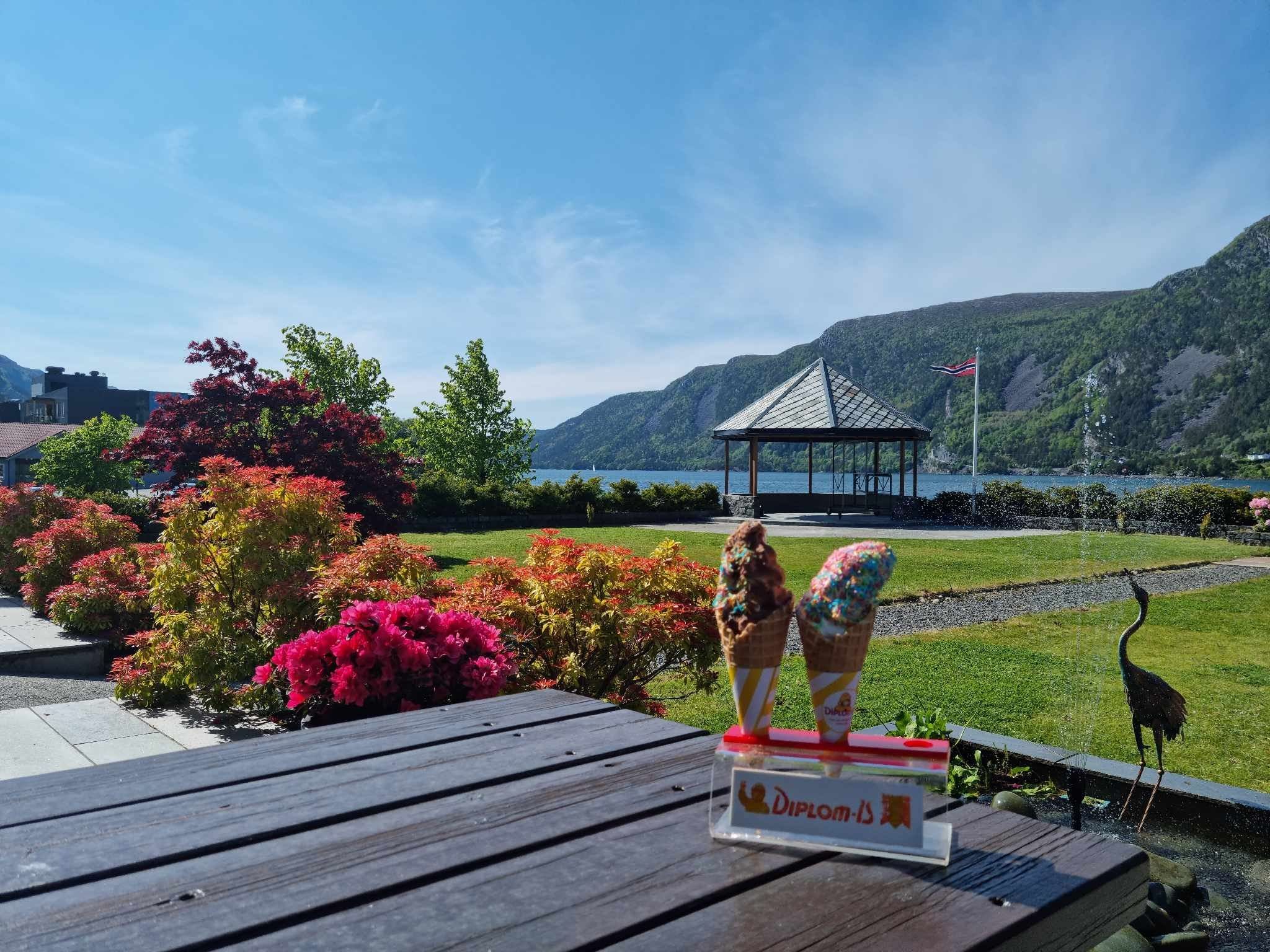 To iskremkjeks på eit bord i forgrunnen, med blomsterhage, lysthus og fjord med fjell under blå himmel på Bremanger Fjordhotell..