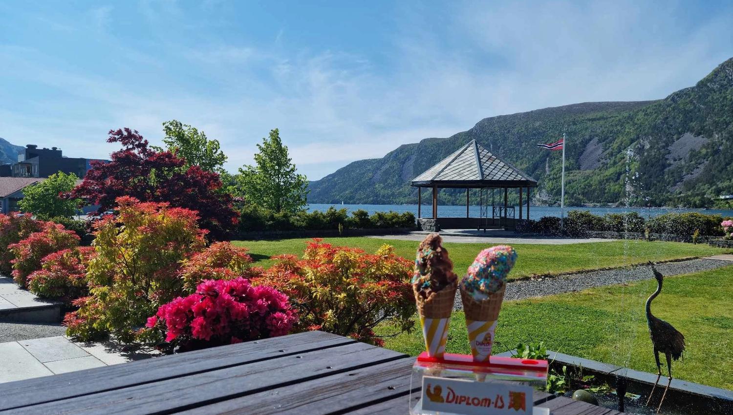 To iskremkjeks på eit bord i forgrunnen, med blomsterhage, lysthus og fjord med fjell under blå himmel på Bremanger Fjordhotell..