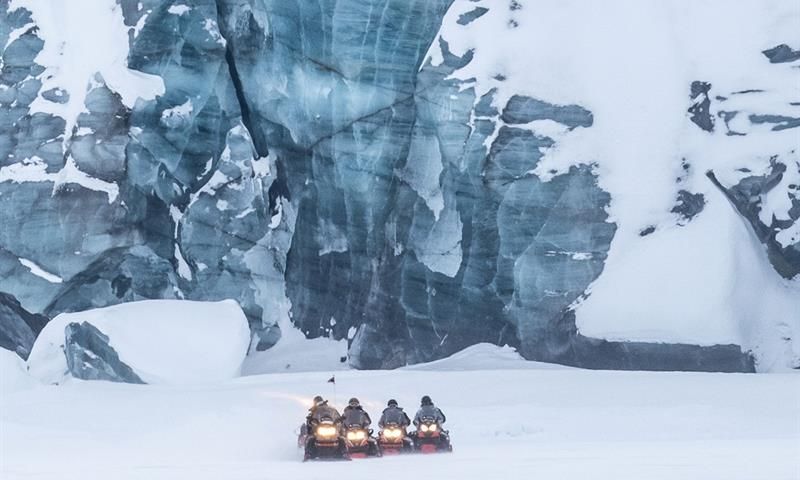 Snøscootere ved foten av en høy isvegg.