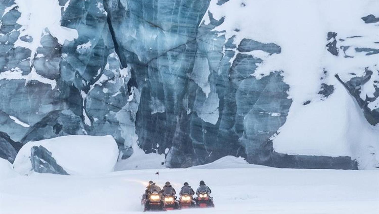 Snøscootere ved foten av en høy isvegg.
