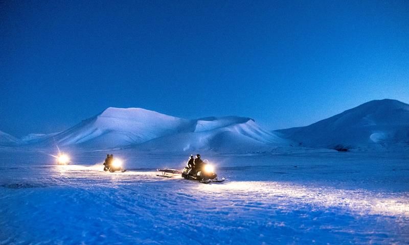 Tre snøscootere kjører på snøen med fjell i bakgrunnen og mørkeblå nattehimmel