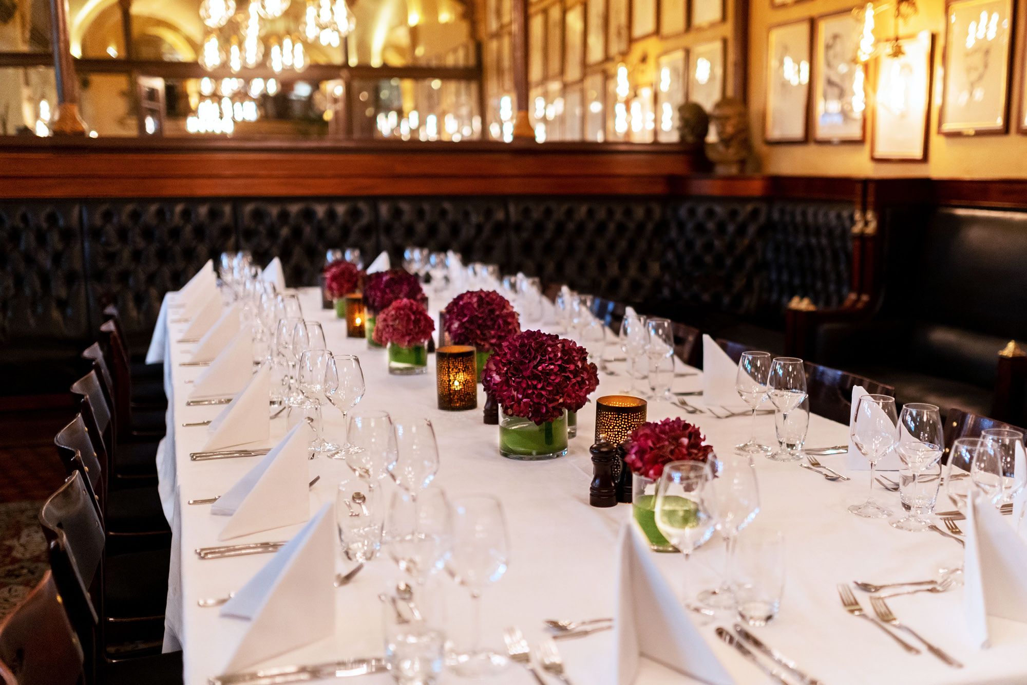 A long table with a white tablecloth and red flowers on, set for dining.